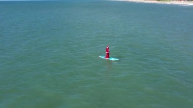 Sörf üzerinde hediye paketi olan Noel Baba. Noel Baba denizde sörf tahtasında. Adam Noel Baba kostümüyle spor yapıyor. Noel ve Yeni Yıl konsepti. Noel Baba 'ya mavi ve yeşil okyanusta kürek ve hediye çantasıyla havadan izleyeceğiz.