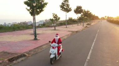 Beyaz motosikletli Noel Baba. Hediye kutusu ve gün batımında hediye paketi. Yeni yıl ve Noel içeriği. Noel Baba kostümlü çocuk bisikletle bize geliyor. Havadan yuvarlak görünüm 