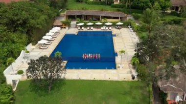 Beyaz ve kırmızı mayo giymiş seksi kızlar havuzda gülümsüyor, açık güneşli yaz havası var. Güzel kızlar lüks bir tropik otelin havuzunda oturuyor, el sallıyor, rahatlıyor, insansız hava aracı yüksek görüşlü.