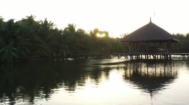 İnsansız hava aracı gölet üzerinde alçaktan uçar, köprünün üzerinden uçar ve palmiye ağaçlarının üzerinden günbatımına doğru uçar.