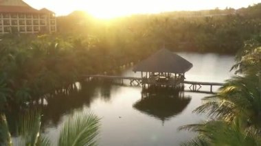 Göletteki tropikal kulübeye hava görüntüsü. Drone gün batımında bir sürü palmiye ağacıyla birlikte göletin üzerinde uçar. Altın arka ışık.
