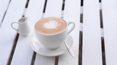 Beyaz kapuçino bardağı, üzerinde kalp resmi olan beyaz ahşap masa. Masanın üzerinde beyaz bardakta kahve 