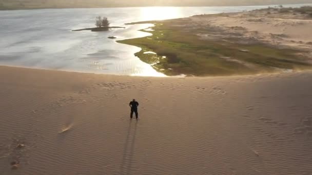 日落时 身穿黑色衣服的老人在沙丘上练习功夫 大量的沙子 金色的落日光芒 运动训练 英俊的男人在沉思 从空中环视沙漠和人类 — 图库视频影像