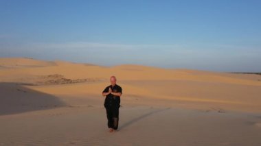 Siyah giyen yaşlı adam gün batımında kum tepelerinde kung fu yapıyor. Bir sürü kum, altın gün batımı ışığı. Spor eğitimi. Yakışıklı adam meditasyon yapıyor. Hava manzarası, insansız hava aracı manzarası