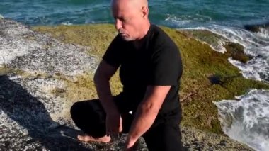Bir kung fu ustasının yakın çekimi, deniz kıyısında pratik yapıyor, elleriyle senkronize nefes egzersizleri yapıyor. Deniz dalgaları onun yanında. Okyanus kıyısındaki taş kıyısı