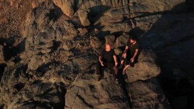 İki adam kayaya oturup konuşuyor. Gün batımında deniz kenarında insanları eğlendirmek için hava manzarası. Altın ışık, mavi gökyüzü ve okyanus