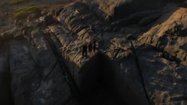 Üç kişi taştan sahilde, siyah çamaşırların içinde bekliyor. Gün batımında okyanus kıyısında spor yapmak için hava manzarası. Altın ışık, mavi gökyüzü ve okyanus. İnsansız hava aracı uçuyor