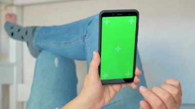 Yeşil ekran ve krom anahtarı olan bir telefon. Rahatlama konsepti. Green Mock-up Screen ile Smartphone kullanarak Kanepede uzanan, Swiping, Scrolling Jestleri yapan kadın. Cep Telefonu Kullanan Kız