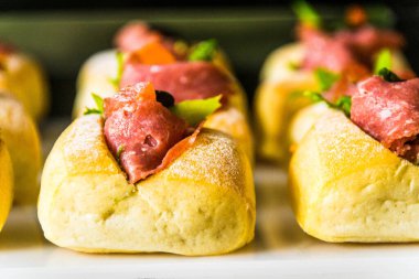 Beyaz tepside servis edilen jambonlu, zeytinli, marullu mini sandviçler.