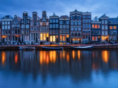 Iconic Amsterdam kanalları ve alacakaranlıktaki geleneksel Hollanda mimarisi. Sıcak şehir ışıkları suya yansıyor. Alacakaranlıktaki Amsterdam tarihi bölgesinin akşam manzarası. Parlayan camlar ve sakin su yansımaları var..