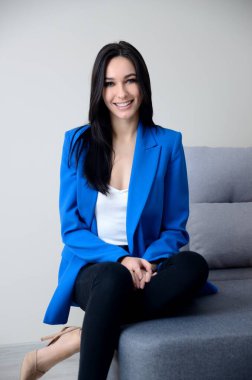 Confident, young woman with blue jacket sits on grey sofa. Beautiful brunette, Miss Lower Silesia poses at home, indoor photo session. 