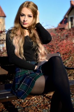 Charming young woman with blonde hair extension sitting on bank in small town. Polish model with black tights, short skirt and black top. clipart