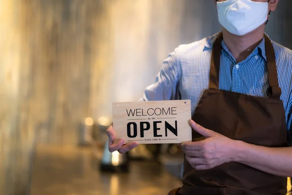 Coffee shop owner with face mask holding holding open sign after lock down and re-open again.