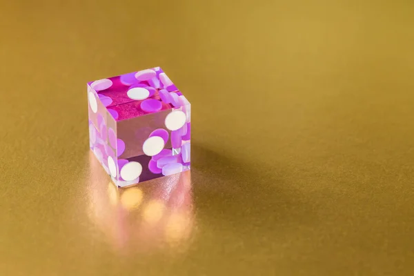 One glassy pink dice on a gold background in sunlight. The result is one. Selective focus macro photography.