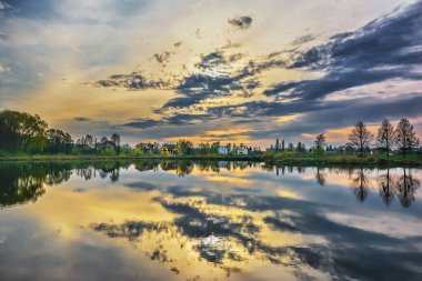 Göl kıyısında gün batımı. Gökyüzü bulutlarla, ağaçlarla ve evlerle suya yansıyor..