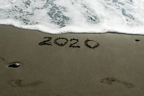 Islak kumsalda 2020 numara, iki ayak izi ve her şeyi silmek üzere olan köpüklü bir dalga.