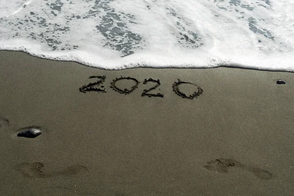 一个写在湿沙滩上的2020年的数字 两个脚印和一个即将抹去一切的泡沫波 图库图片