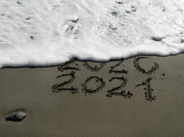2020年和2021年的数字写在湿沙滩和一个泡沫般的波浪上 这将抹去这一信息 免版税图库照片
