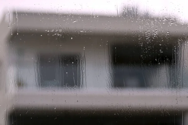 Wet window glass with raindrops, water drippings and a pale house shape