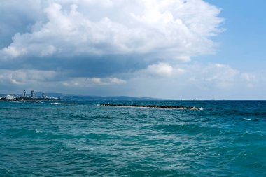 Dalgalı deniz suyu yüzeyi, mavi gökyüzü fırtınalı bulutlar, dağlar, kıyı şeridi ve ufukta gemiler.