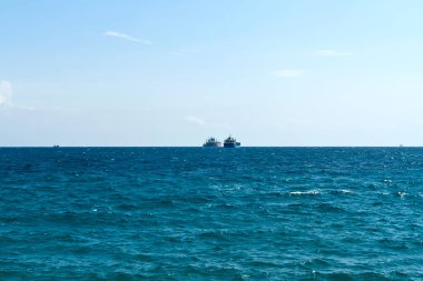 Dalgalanan deniz suyu yüzeyi, açık mavi gökyüzü ve ufuktaki gemiler.