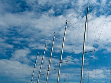 Mavi gökyüzü ve beyaz bulutların önünde metal yat direkleri.