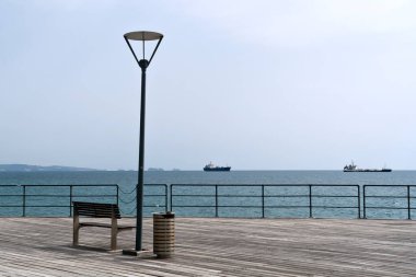 Boş bank ve sokak lambası, denizin, gökyüzünün ve ufuktaki gemilerin önündeki ahşap iskelede, Limasol, Kıbrıs