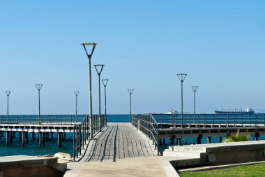 Açık mavi gökyüzü ve ufukta gemilerin önünde sokak lambaları olan boş ahşap iskele.