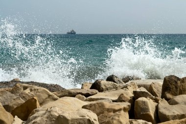 Büyük beyaz kayalara vuran deniz dalgaları, ufukta deniz manzarası ve gemi.