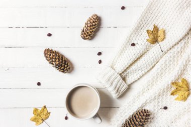 Sonbahar veya kış kompozisyonu. Bir fincan kahve, kadın süveteri, kozalak ve beyaz ahşap arka planda düşen yapraklar. Düz konum, üst görünüm, kopyalama alanı.