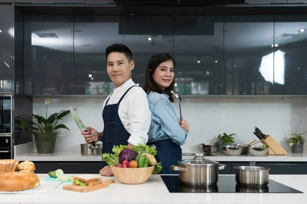 Kendine güvenen aşçı ve kadın bildirileri yemek pişirme mutfağında bir çorba kepçesiyle bıçak tutarak sırt sırta dururlar. Önlüklü Asyalı çift hafta sonları sağlıklı yemek pişirmeye hazırlanıyor..