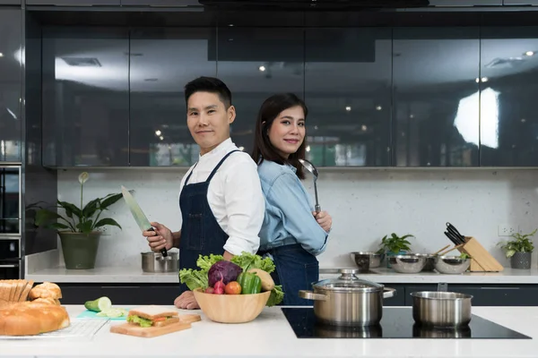Kendine güvenen aşçı ve kadın bildirileri yemek pişirme mutfağında bir çorba kepçesiyle bıçak tutarak sırt sırta dururlar. Önlüklü Asyalı çift hafta sonları sağlıklı yemek pişirmeye hazırlanıyor..