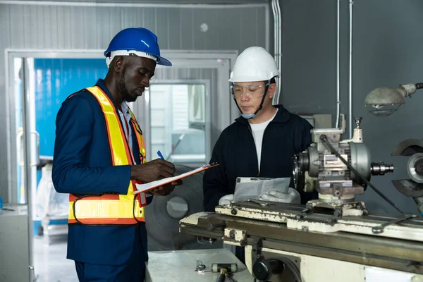 Afrikalı Amerikalı endüstriyel mühendis Asyalı işçilere tavsiye vermek için kontrol makinesini kullanıyor. İş gününde imalat fabrikasında çalışan çok ırklı teknisyen. Irk eşitliği kavramları.