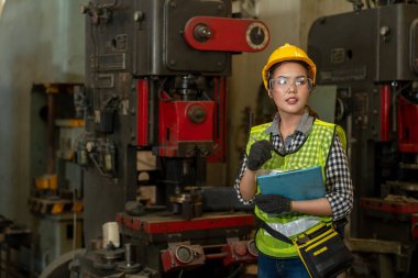 İş gününde fabrikada makineleri kontrol ederken elinde pano tutan Asyalı kadın teknisyen. Partikül solunum maskesi takan kendine güvenen bir işçi. Endüstriyel üretim