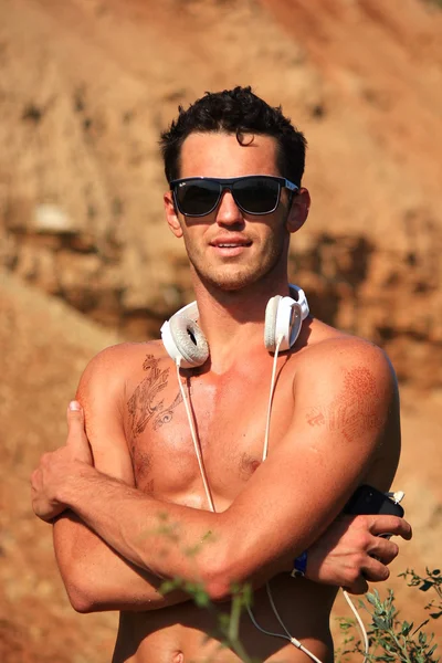 Серйозний голий м'язистий молодий чоловік, що стоїть у пустелі, посміхається, прийнявши себе. Демонструє спортивну фігуру, м'язи, слухає музику. Носіння сонцезахисних окулярів та білих навушників — стокове фото