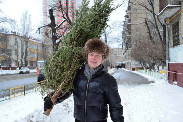 A young guy in fur cap carries a Christmas tree home on the street — Stock Photo, Image