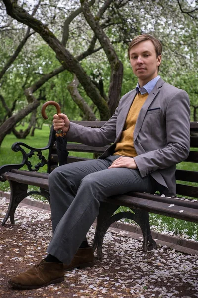 Een jonge man in een grijs pak met een zwarte paraplu-stok op de Bank in de weelderige appelboomgaard — Stockfoto