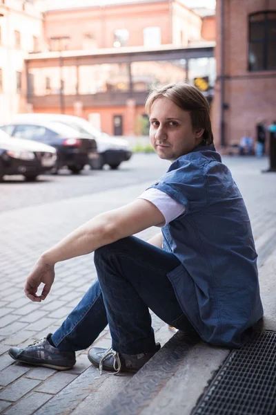 The young man sits on the stairs and smiles — Stock Photo, Image