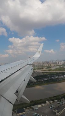 VERTICAL FOOTAGE, Yolcu uçağı alçaktan inmek üzere, pencerenin kanatları mavi gökyüzü ve beyaz bulutlar, ve yerde yüksek binalar var