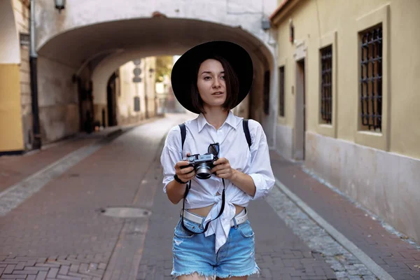 若い女の子写真家 その少女は黒い髪をしている 彼女は黒い帽子とサングラスをかけていて カメラは古いヨーロッパの街の道路に立っています — ストック写真