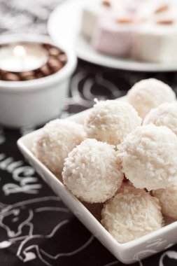 Siyah masa örtüsünün üzerindeki kasede birkaç raffaello var. Kahve çekirdeği dolu cam kavanoz. Üstünde mum var. Arka plan kadar tatlı.