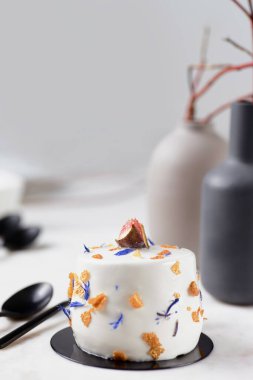 Mermer bir tahtanın üstünde incir olan beyaz pasta. Bir mermer tahtada iki çay kaşığı. Arka planda iki vazo var. Arkaplan beyaz.