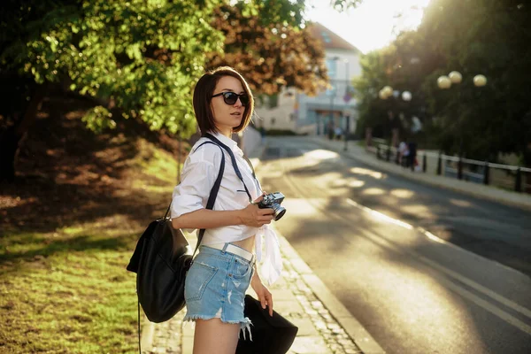 Девушка в черной шляпе и солнцезащитных очках с фотокамерой на дороге в старом европейском городе. — стоковое фото
