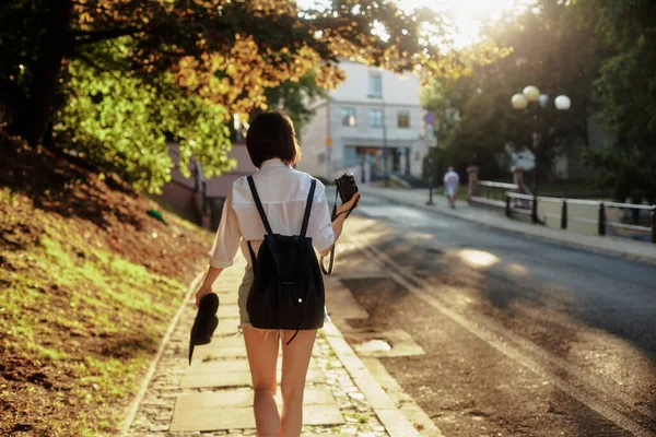 La fille au chapeau noir et lunettes de soleil avec appareil photo sur une route dans une vieille ville européenne. — Photo
