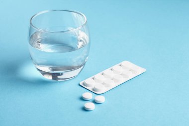 Mavi arka planda bir bardak su ve su kabarcığı vardı. Beyaz haplar paketten dökülüyor..