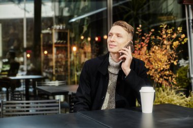 Caddedeki bir kafede oturan bir adam elinde bir fincan kahveyle telefonda konuşuyor. Sonbahar