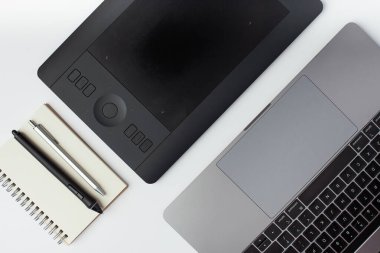 Üst görünüm fotoğrafı, bir not defteri, kalem, siyah grafik tasarım tableti, stilus ve beyaz zemin üzerinde bir laptop.