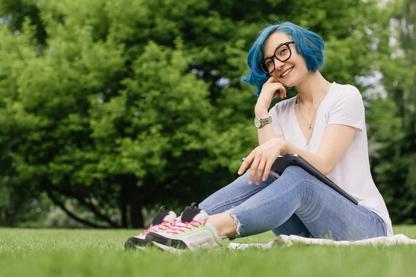 Mavi saçlı genç kadınlar ellerinde dizüstü bilgisayarlarıyla yeşil çimlerde oturuyorlar. Yaz zamanı.