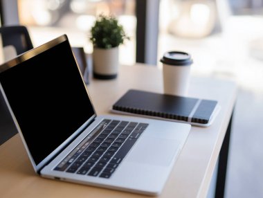 Bir masa, doğal ışık alan parlak bir çalışma alanında dizüstü bilgisayar, defter ve kahve fincanı içerir..