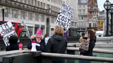 10 Mart 2020: Protesto plaklarıyla grev yapan öğretim üyeleri ve öğrenciler arka planda kırmızı çift katlı otobüsler geçerken grev hattına gülüyorlar